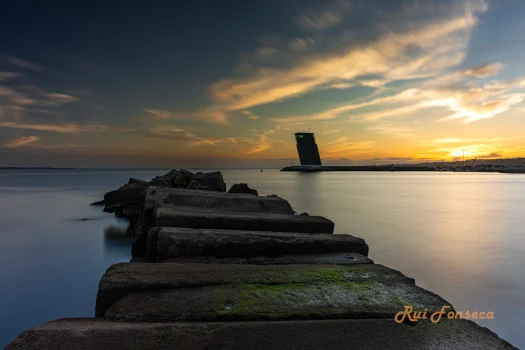 A long exposure photograph of a stone pier extending into the sea at sunset, with the Leuchtturm Helgoland lighthouse in the background.