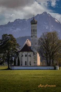 A picturesque view of a church with a distinctive onion-domed bell tower, set against a backdrop of snow-capped mountains and a lush green meadow.