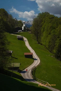 A lone figure in a red jacket walks along a winding road in a picturesque alpine landscape. The road curves gently uphill, flanked by lush green meadows and traditional wooden structures. In the distance, snow-capped mountains rise majestically against a clear blue sky. The scene evokes a sense of tranquility and natural beauty.