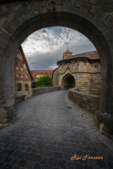 A cobblestone path leads through an arched stone gate in a medieval town, with traditional half-timbered houses and stone buildings visible on either side. The sky is overcast.