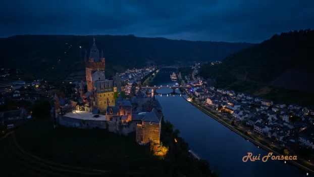 An aerial view of a castle illuminated at night, overlooking a river and a town.
