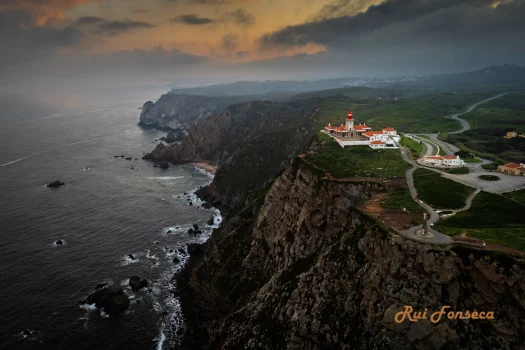 A dramatic aerial view of the Cabo da Roca lighthouse in Portugal, with a stormy sky and rugged coastline.