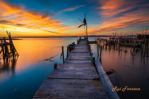 A wooden pier extends into a calm body of water under a vibrant sunset sky.