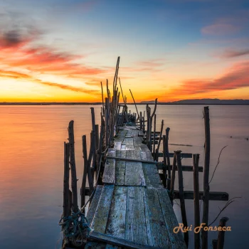 A weathered wooden pier extends into a calm body of water under a vibrant sunset sky.