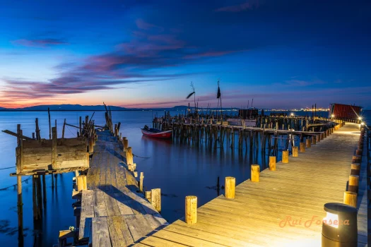 A long wooden pier extends into a calm body of water at dusk, with a colorful sunset reflecting on the water's surface. A small fishing boat is moored near the end of the pier.
