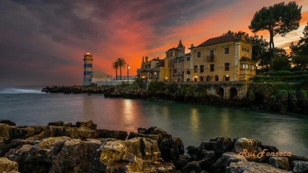 A picturesque coastal scene featuring a historic villa and a lighthouse at dusk, with a vibrant sunset sky.