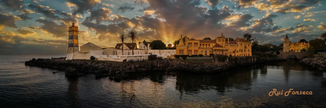 Panoramic view of a lighthouse and a grand building complex on a rocky peninsula jutting into the sea, bathed in the warm light of a sunset.