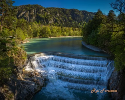 A cascading waterfall flows into a turquoise river, framed by lush green mountains and dense vegetation.