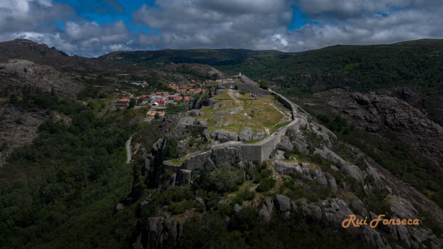 Aerial view of Castelo Lindoso, a medieval castle ruin perched atop a hill in the Minho region of North Portugal, surrounded by a rural landscape of heather, grassland, and scattered deciduous trees. The sky is partly cloudy, creating a dramatic and serene atmosphere.