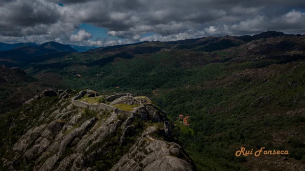 A dramatic aerial view of Castelo Lindoso, a medieval castle perched atop a rocky outcrop in northern Portugal. The castle walls and stone structures are visible against a backdrop of lush green valleys and rolling mountains under a partly cloudy sky.