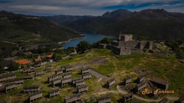 Aerial view of Castelo Lindoso, a medieval castle in Portugal, surrounded by traditional stone huts and a scenic landscape with mountains and a lake.