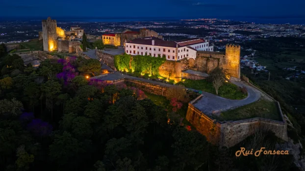 Aerial view of Guimarães Castle and Palace of the Dukes of Braganza illuminated at night, with a town visible in the background.