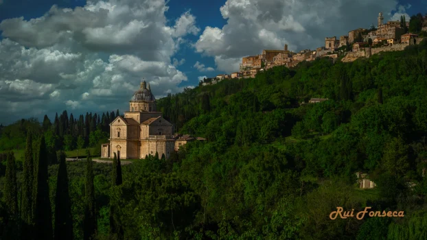 A picturesque view of a Renaissance-era church and a hilltop town in Italy, bathed in soft light and framed by lush greenery and a dramatic sky.