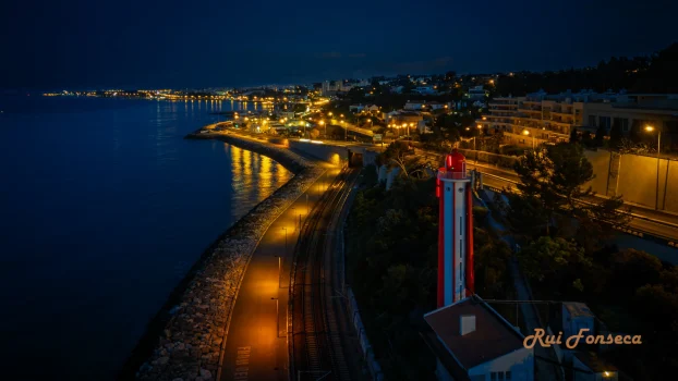 Aerial view of a coastal city at night, with a distinctive red and white lighthouse in the foreground.