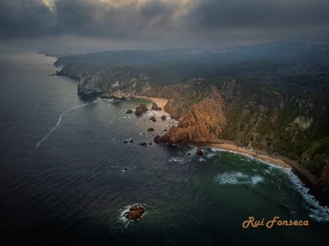 Aerial view of a rugged coastline with dramatic cliffs, a sandy beach, and a turbulent sea under a stormy sky.