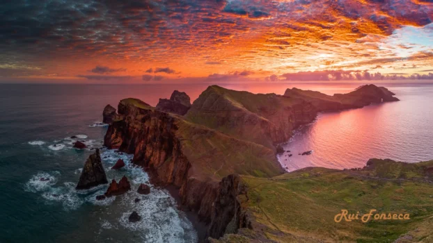 Aerial view of dramatic cliffs and coastline at sunset, with vibrant orange and pink skies reflecting on the water.