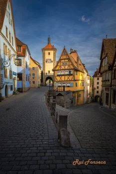 A cobblestone street leads towards a medieval gate in a historic European town, framed by traditional timber-framed buildings.