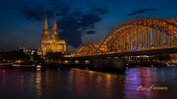 Night view of the Rhine River with the Cologne Cathedral and Hohenzollern Bridge in the background.