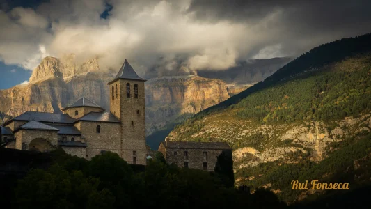 A dramatic, low-angle photograph of a stone monastery nestled in a valley, with towering mountains and stormy skies in the background.