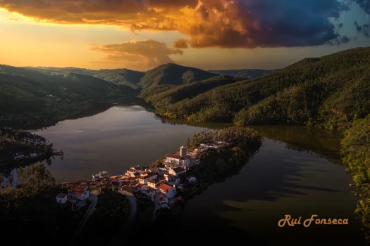 Aerial view of a white-washed village nestled on a peninsula jutting into a wide, calm river, surrounded by lush green hills.