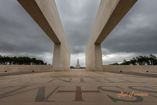 A low-angle perspective shot through monumental stone pillars frames a distant view of a church spire under a cloudy sky. The foreground features a paved area with a large, stylized cross inlaid in the stone. The overall scene evokes a sense of solemnity and historical significance.