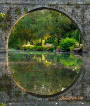 A symmetrical photograph of a stone archway framing a lush green landscape reflected in calm water, creating a circular composition.