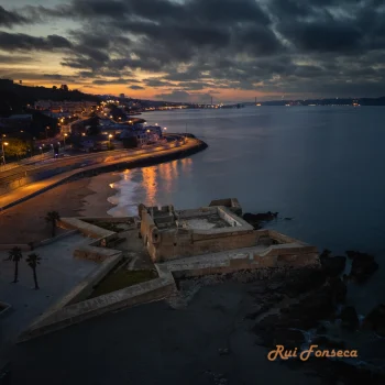Aerial view of a coastal fortress at dusk, with a city skyline visible in the distance.