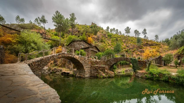 A picturesque stone bridge arches gracefully over a tranquil river, surrounded by lush woodland and a dramatic, overcast sky. The scene evokes a sense of history and natural beauty.