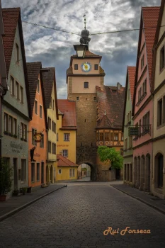 A cobblestone street leads towards a medieval tower in a historic European town. Buildings with traditional architecture line the street, and the sky is overcast.
