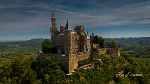 Aerial view of Hohenschwangau Castle in Bavaria, Germany, surrounded by lush greenery and a clear blue sky.