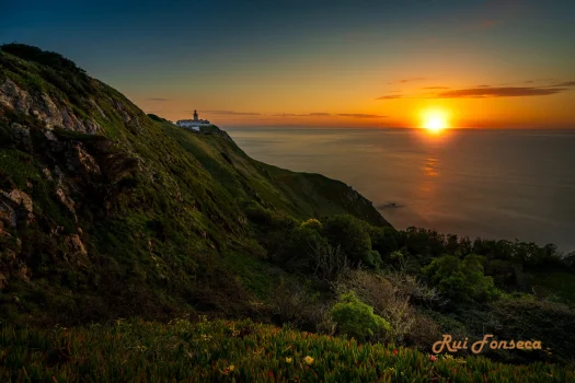 A scenic view of a lighthouse perched atop a cliff overlooking the ocean during a vibrant sunset.