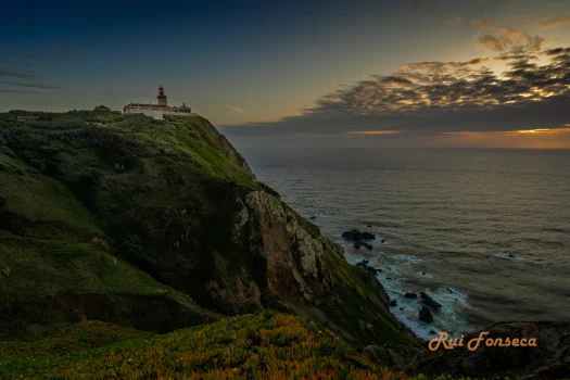 A scenic coastal landscape featuring a lighthouse perched atop a dramatic cliff overlooking the ocean at sunset.