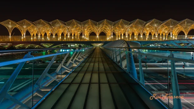 Nighttime long exposure photograph of a pedestrian bridge leading to the distinctive roof of Lisbon Airport.