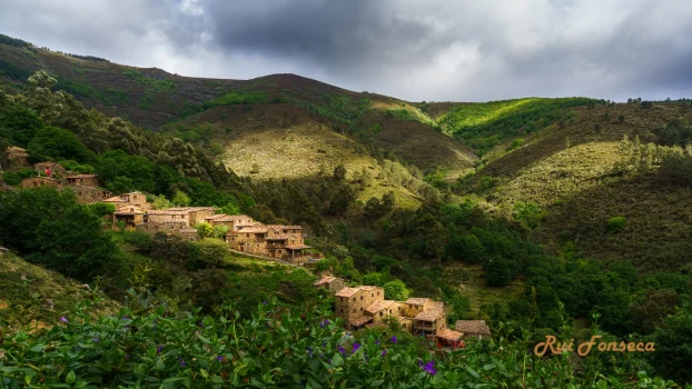 A picturesque village nestled in a lush, green valley, framed by rolling hills under a dramatic, cloudy sky.
