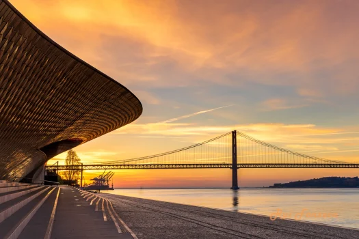 A stunning photograph captures a vibrant sunset over a cityscape, featuring a modern architectural structure in the foreground and a prominent suspension bridge in the background. The scene is bathed in warm, golden light, creating a picturesque and serene atmosphere.