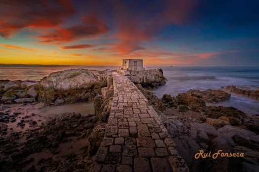 A picturesque coastal scene at sunset featuring a stone causeway leading to a small, white building perched on a rocky outcrop. The sky is ablaze with vibrant colors, and the ocean gently laps against the shore.
