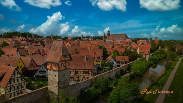 Aerial view of a historic town with distinctive red-tiled roofs, surrounded by a stone wall and a river.