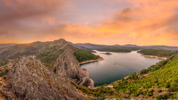 Panoramic view of a lake surrounded by rocky hills and lush green vegetation, with a vibrant sunset sky.
