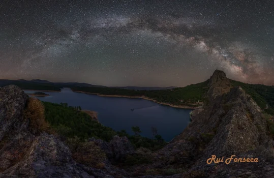 A stunning nightscape photograph showcases a vast, dark lake nestled amidst rocky terrain, with the Milky Way stretching across the sky. The scene is dominated by a prominent rock formation jutting out from the landscape, framing the view of the lake and the celestial display above.