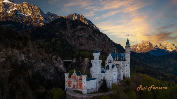 An aerial view of Neuschwanstein Castle nestled amidst a lush green forest and towering snow-capped mountains under a soft, diffused light.