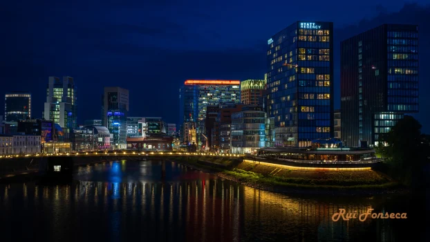 Nighttime view of a modern cityscape reflected in a river, with illuminated buildings and a bridge.
