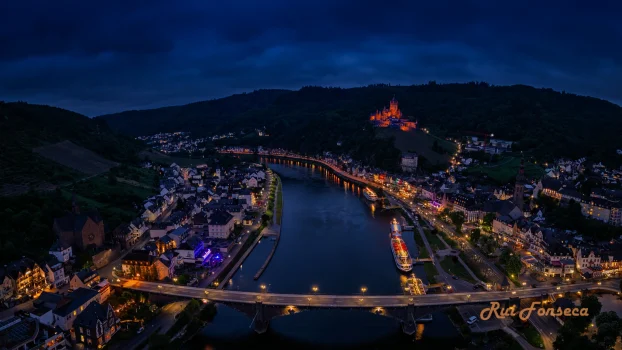 An aerial view of a town at night, with lights illuminating the streets and buildings. A hill rises in the background, covered in trees.