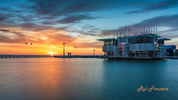 A long exposure photograph of the Oceanário de Lisboa (Lisbon Oceanarium) at sunset, with a vibrant orange and blue sky reflected in the calm water.