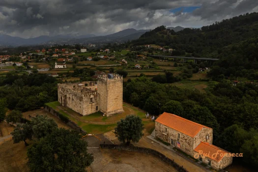 Aerial view of a medieval castle and adjacent building in a rural landscape, with a dramatic sky overhead.