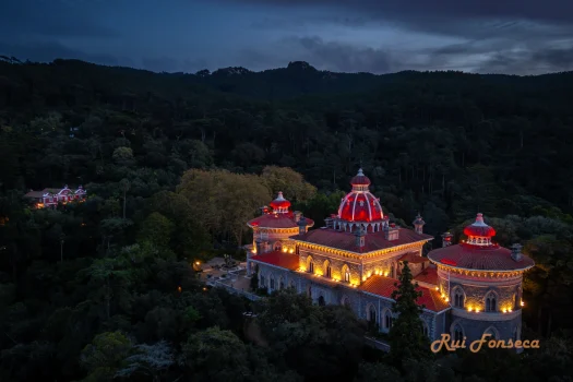 Aerial view of a grand, illuminated palace nestled within a lush, forested landscape at dusk.
