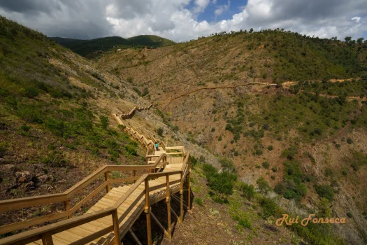 A wooden boardwalk winds down a hillside, offering views of a valley and distant mountains under a cloudy sky.