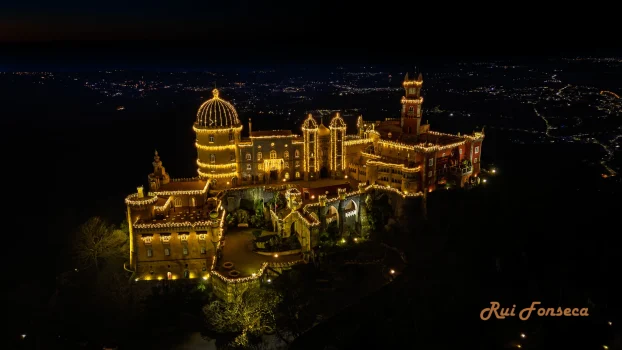 Aerial view of Pena Palace at night, illuminated with warm lights, set against a dark sky with distant city lights.