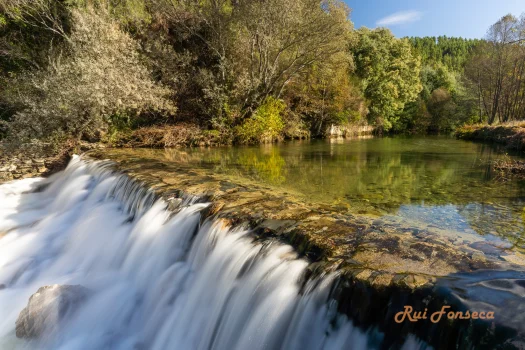 A long exposure photograph of a small waterfall cascading over rocks into a calm, reflective pool of water, surrounded by lush vegetation and trees under a clear blue sky.
