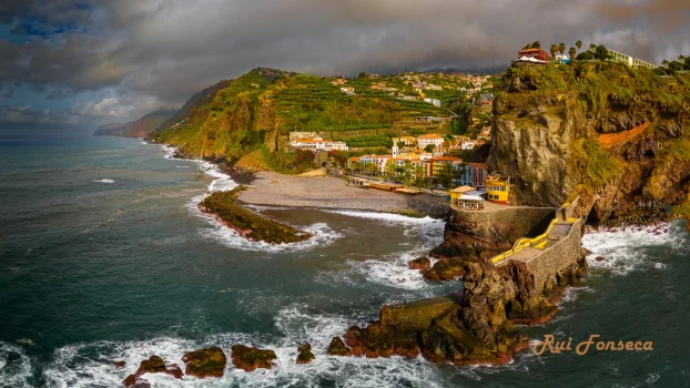 Aerial view of a coastal village nestled into a cliffside, with a beach below and dramatic skies overhead.