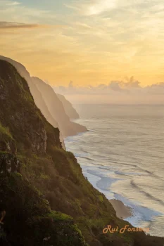 A long exposure photograph of a coastline at sunset, featuring dramatic cliffs covered in lush vegetation, a sandy beach, and the ocean.